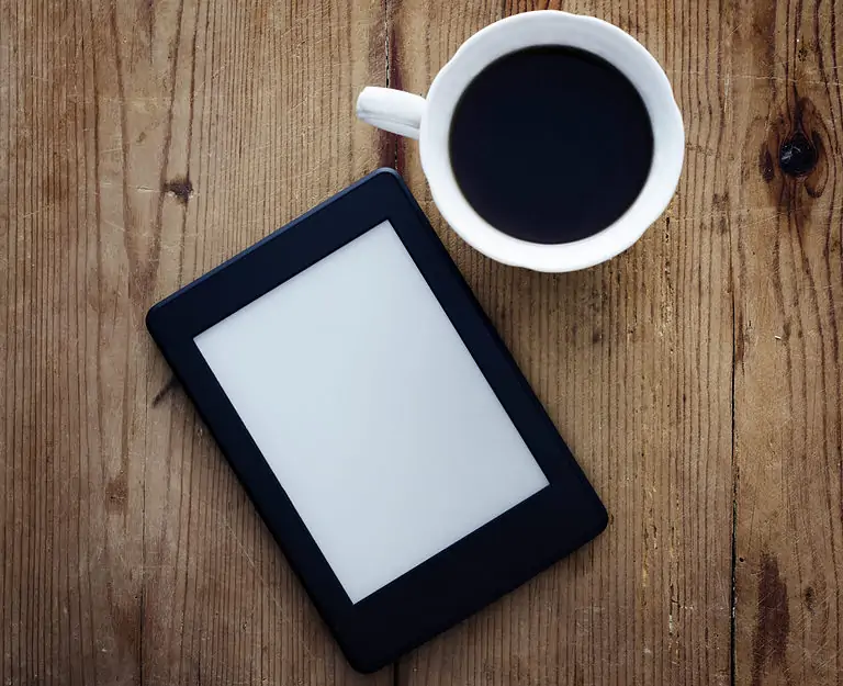 kindle e-reader on wooden table next to a cup of coffee